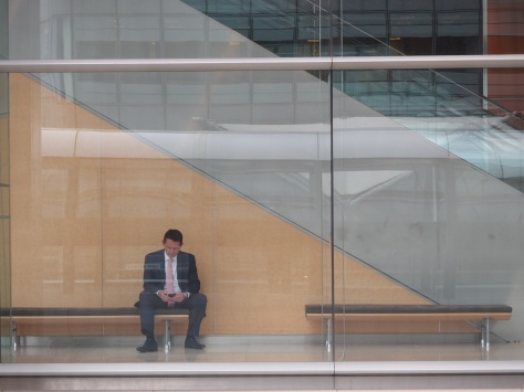 Man sitting on a bench in an office block across from a covered pedestrian walkway at Canary Wharf